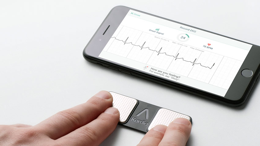 AliveCor monitor with person touching it with 2 fingers from each hand and smartphone showing ECG